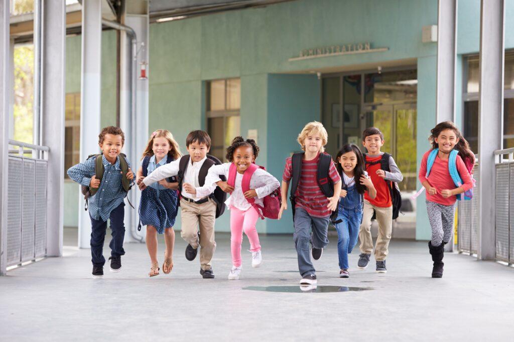 Gruppe fröhlicher Kinder unterschiedlicher Herkunft rennt auf dem Schulgelände einer bilingualen Grundschule in Berlin, voller Energie nach dem Unterricht.