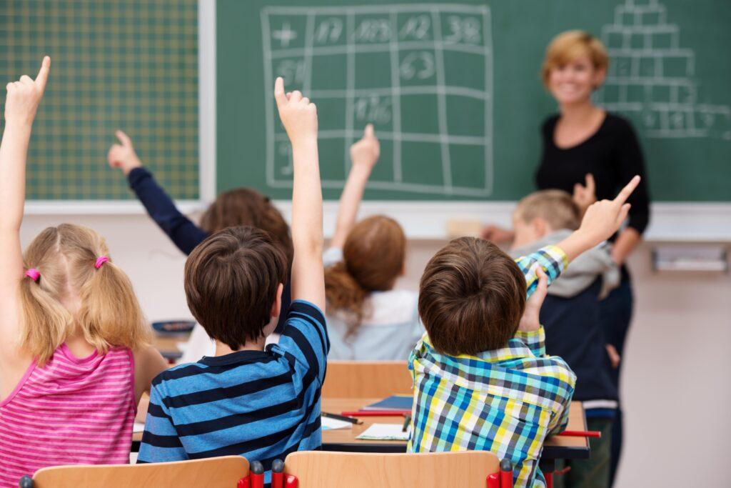 Kinder heben in einem Klassenzimmer einer bilingualen Grundschule in Berlin begeistert die Hände, während eine Lehrerin vor einer Tafel mit mathematischen Aufgaben steht.