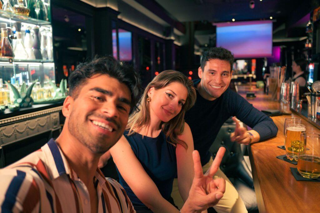 Drei lächelnde Freunde machen ein Selfie in einer belebten Bar in Wien, genießen gemeinsam Bier und die lebhafte Atmosphäre der Lokalität.