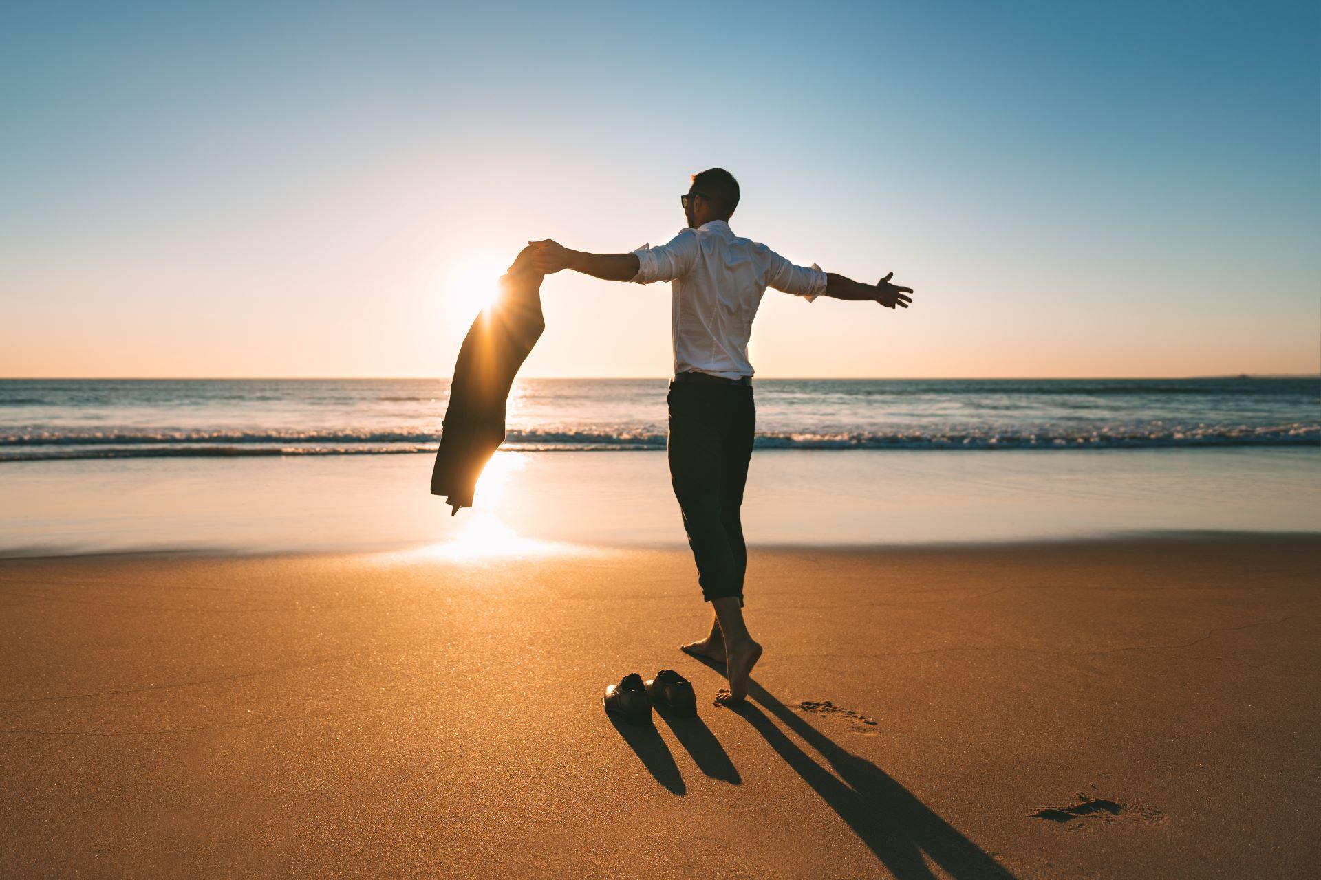 Geschäftsmann am Strand bei Sonnenuntergang, der seine Jacke auszieht und die Freiheit genießt, symbolisch für Work-Life-Balance und Entspannung.