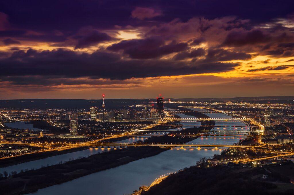 Ein atemberaubendes Nacht-Panorama von Wien mit beleuchteter Skyline, einschließlich der Donau und markanter Hochhäuser unter einem dramatischen Sonnenuntergangshimmel.