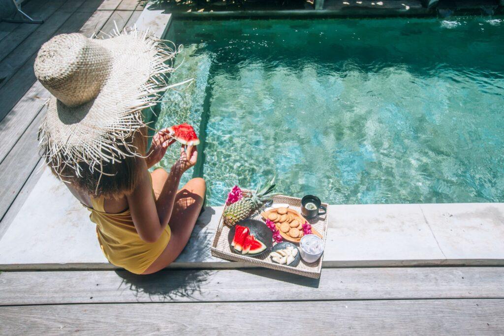 Frau in einem Sonnenhut sitzt am Pool und genießt Bio Snacks wie Wassermelone, Ananas und Kekse. Inspirierende Szene für nachhaltige und gesunde Bio Lebensmittel.