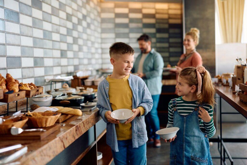 Kinder holen sich Fruehstueck am Buffet in einem Hotel in Kleinarl, waehrend Eltern im Hintergrund den Tag ruhig beginnen